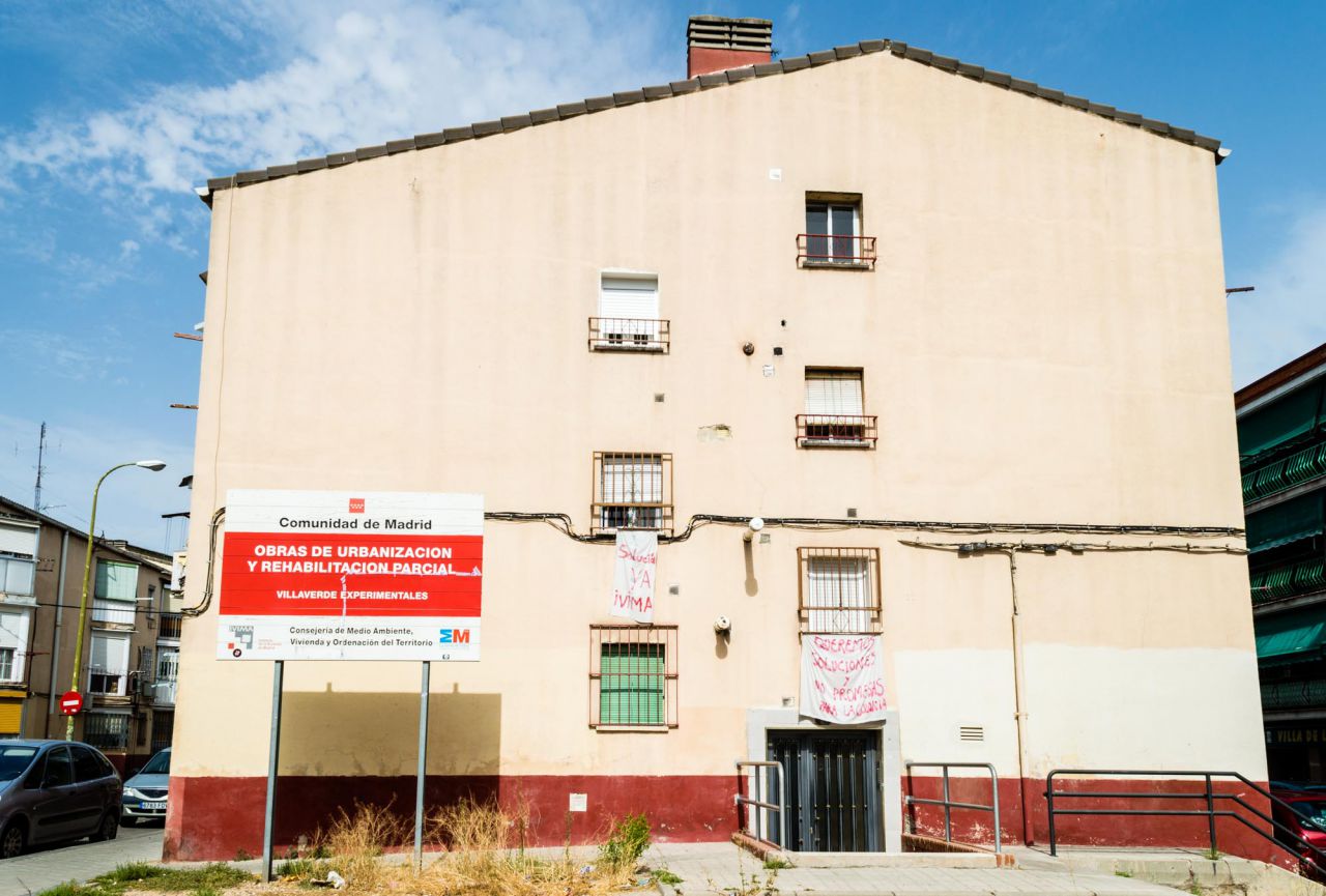 Cartel del programa de rehabilitación enfrente a una fachada con pancartas de protesta.