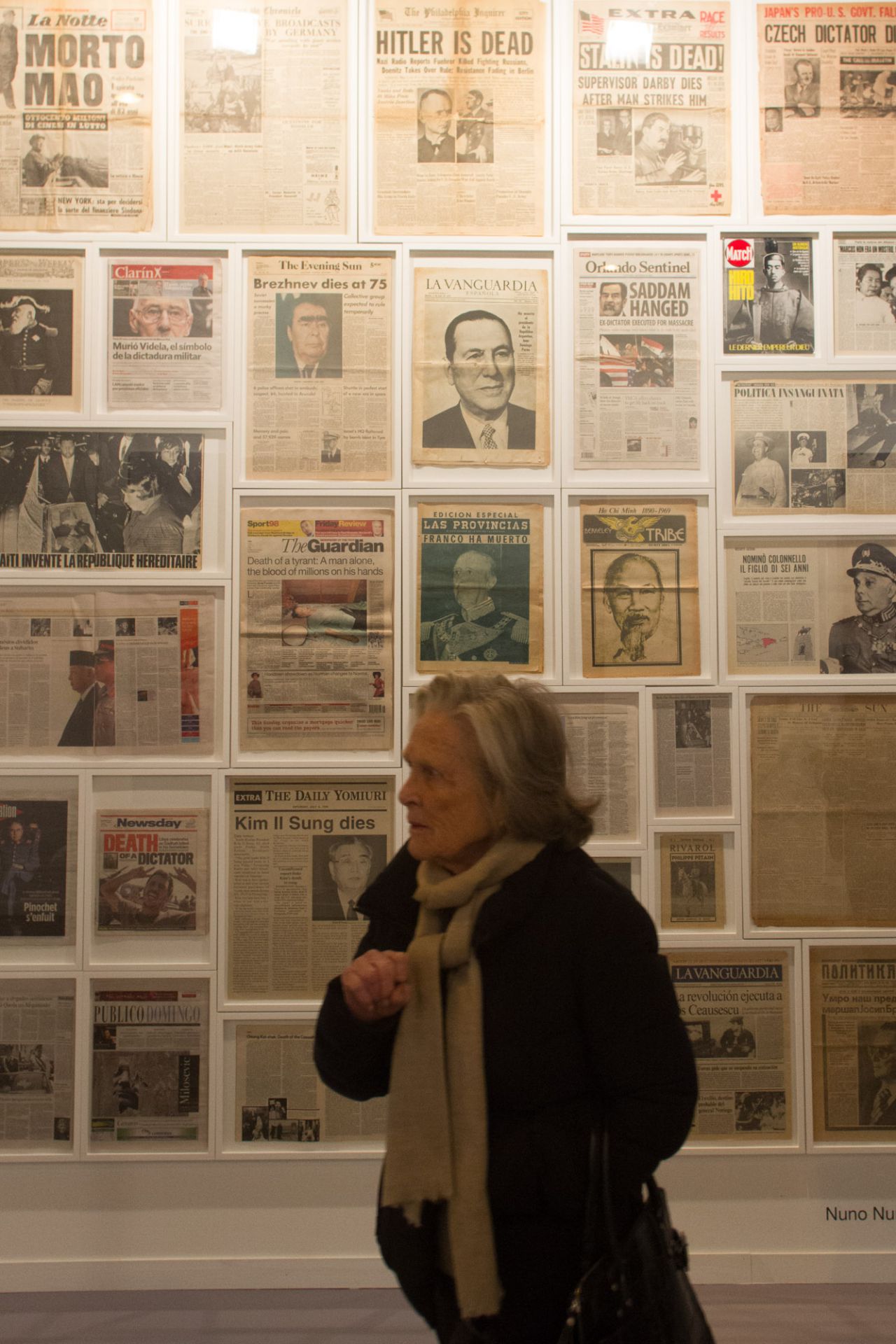 Una mujer pasea frente a una obra hecha con portadas de periódicos que anuncian la muerte de grandes dictadores.